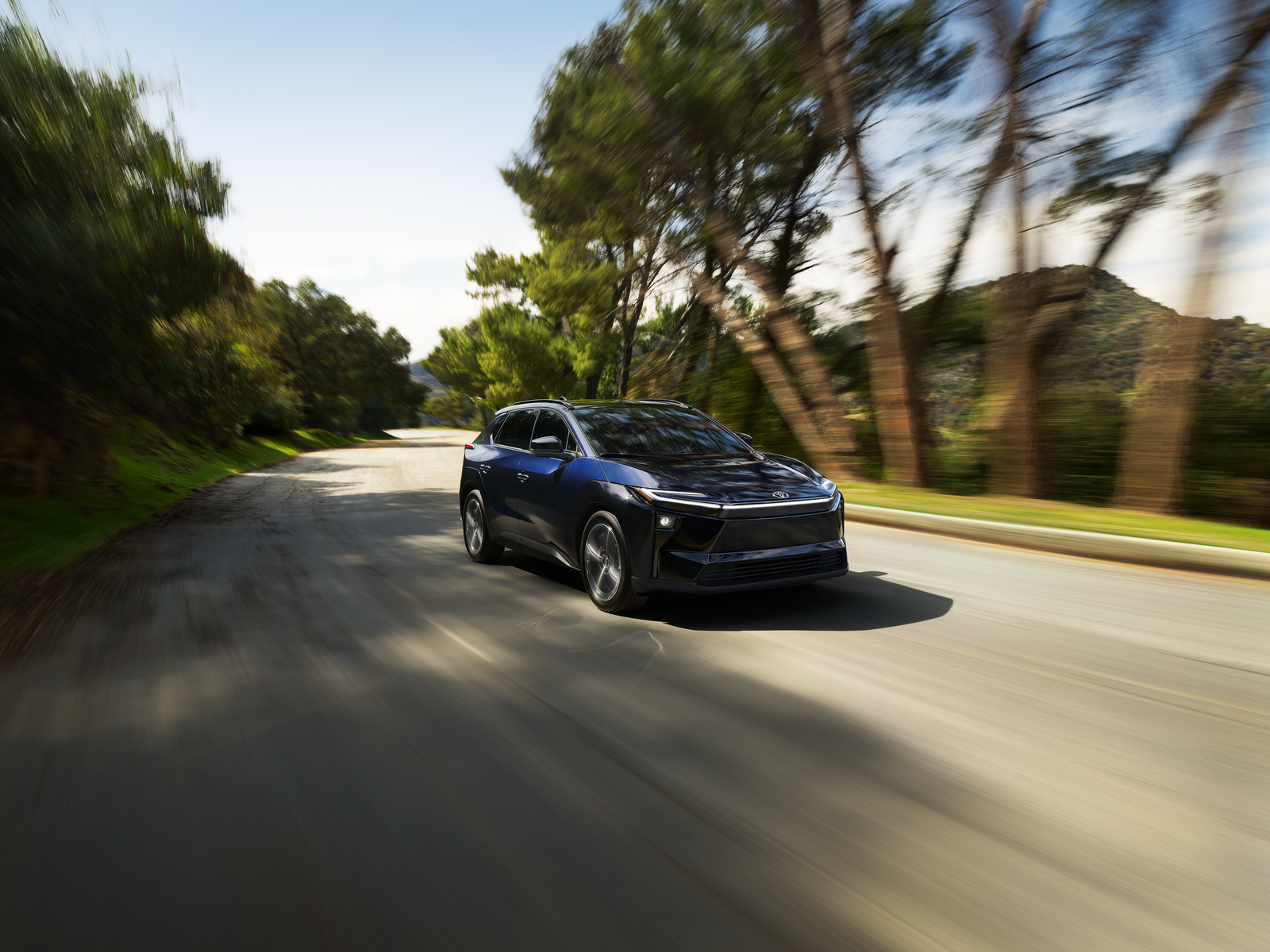Blue Toyota bZ electric SUV driving on a tree-lined road, front three-quarter view