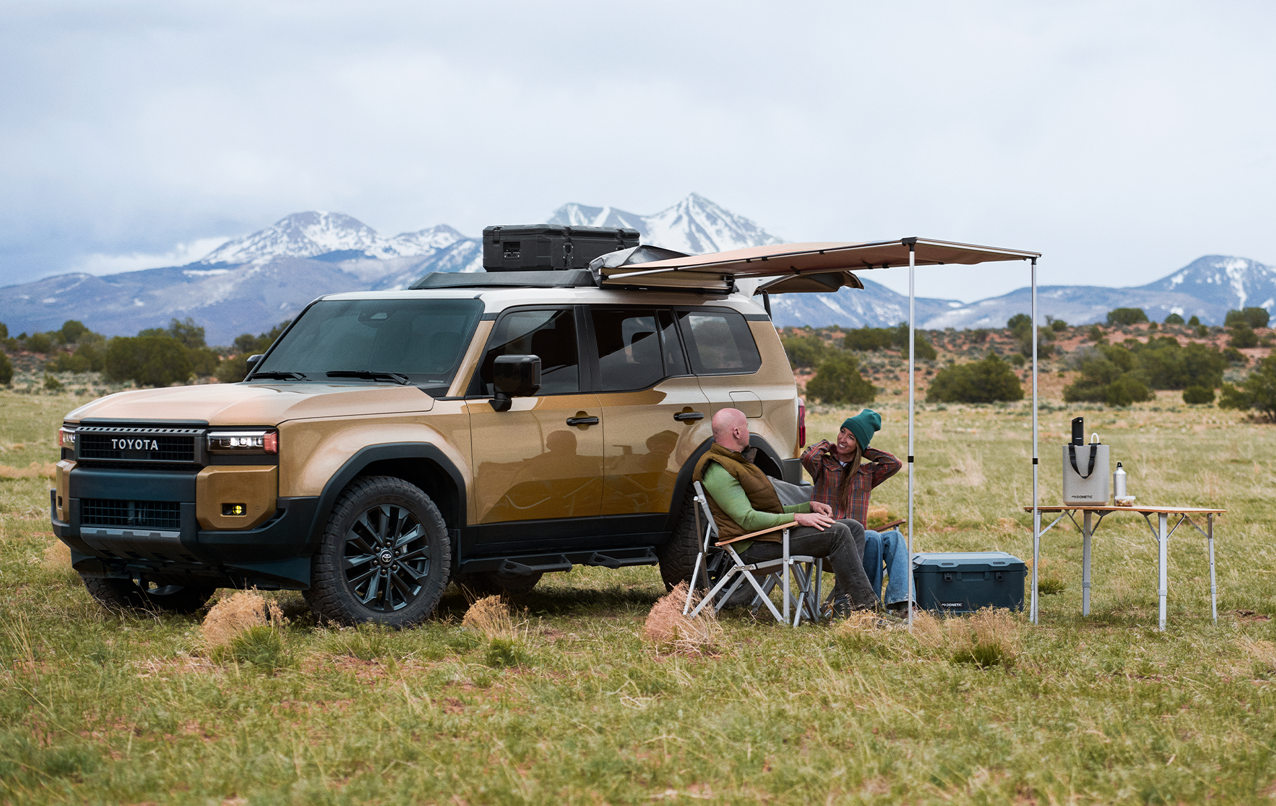 Tan Toyota Land Cruiser with roof box and awning parked at a campsite below snowy mountains